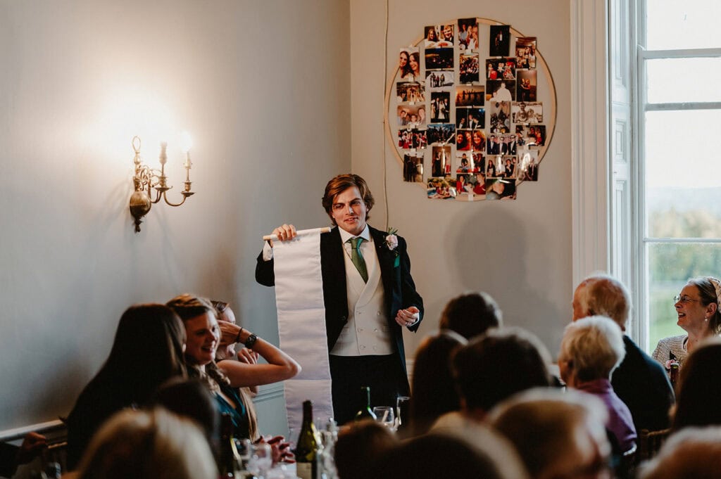 A person in formal attire stands holding a long sheet of paper, addressing an audience in a well-lit room with a collage of photos on the wall behind.
