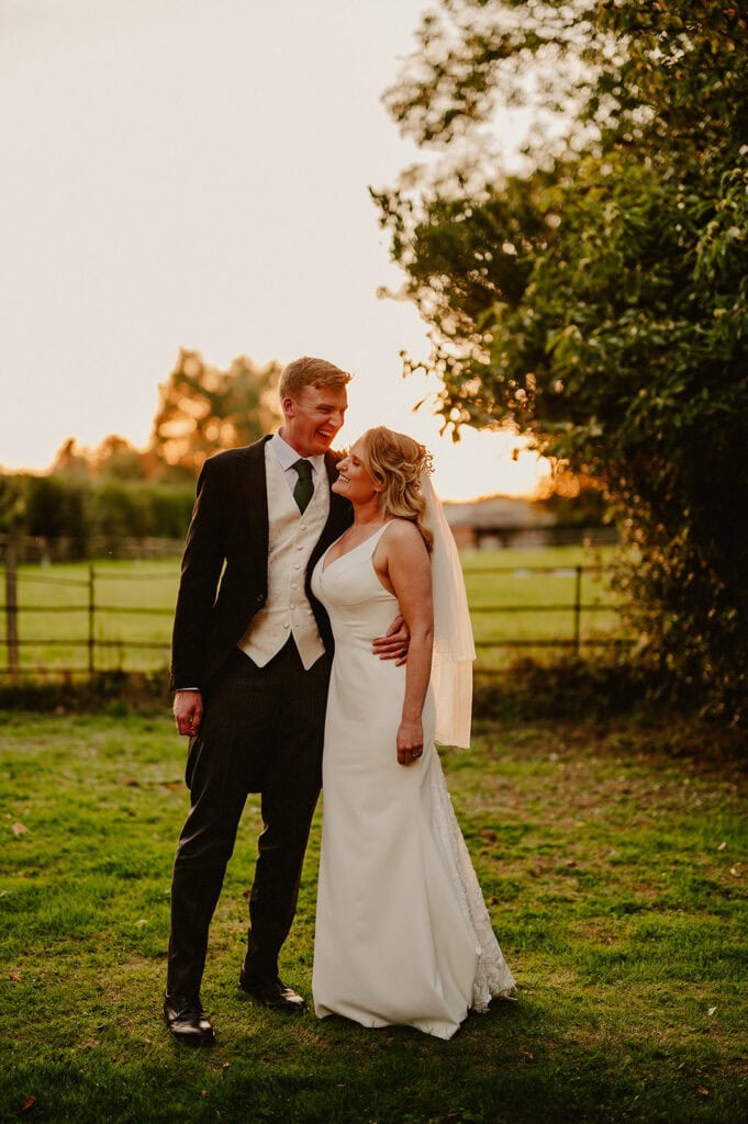 A couple in formal wedding attire stands together on a grassy area at sunset. The groom wears a black suit with a white vest, and the bride wears a white gown with a veil.