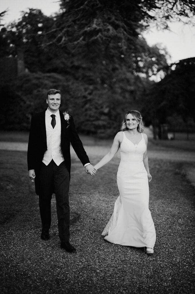 A bride and groom hold hands while walking outdoors. The bride wears a white dress, and the groom is dressed in a black suit with a white waistcoat. Trees and a gravel path are visible behind them.