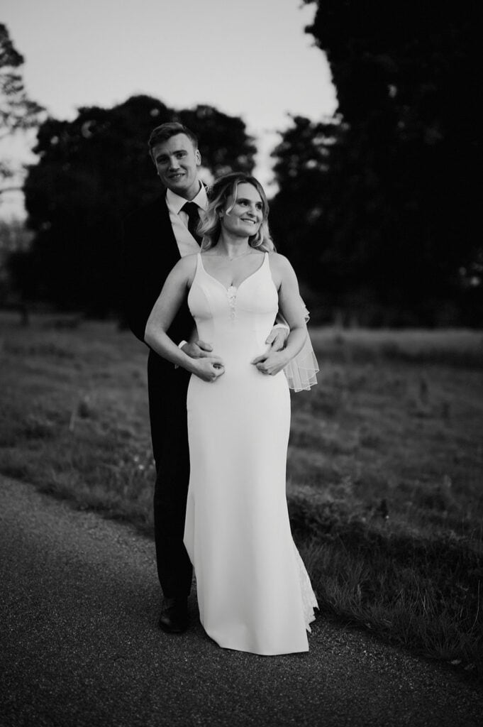 A couple stands on a path, with the man embracing the woman from behind. The woman wears a long white dress, and the man wears a dark suit. Trees and grass are visible in the background.