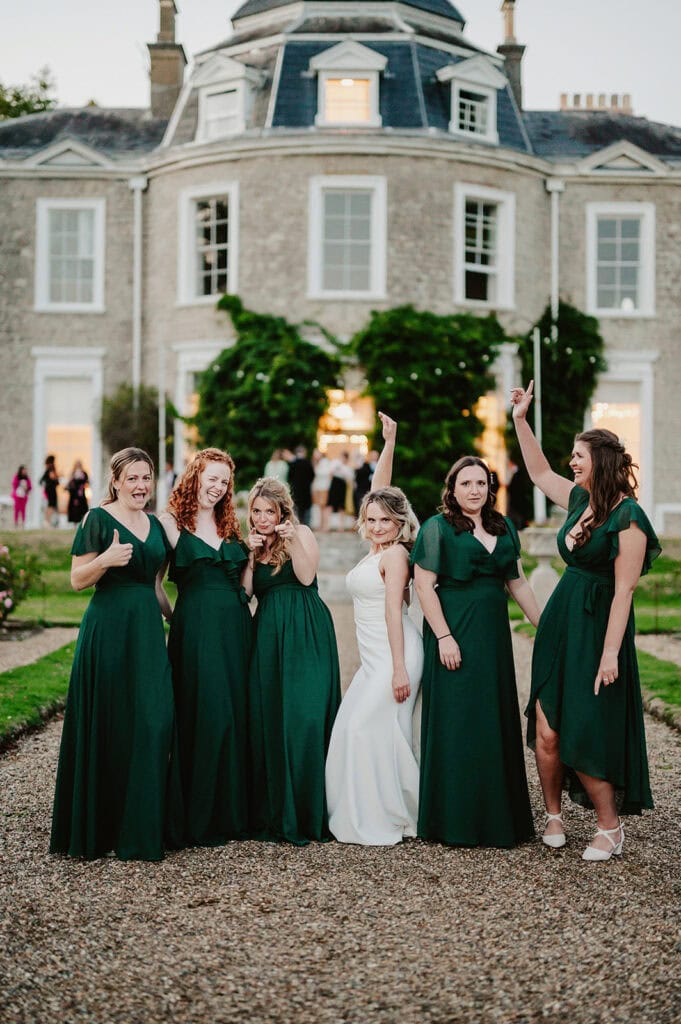A bride in a white dress poses with five bridesmaids in green dresses in front of a large, historic building. The bridesmaids make celebratory gestures.