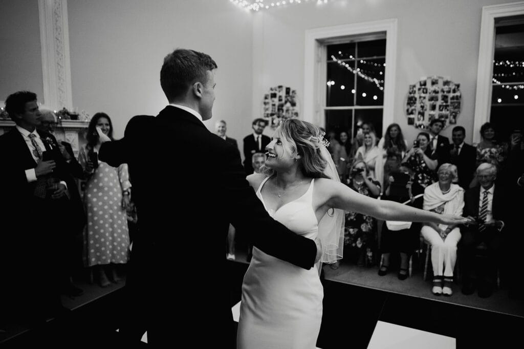 A bride and groom dance at their wedding reception, surrounded by smiling guests seated and standing, with string lights and photo decorations in the background.