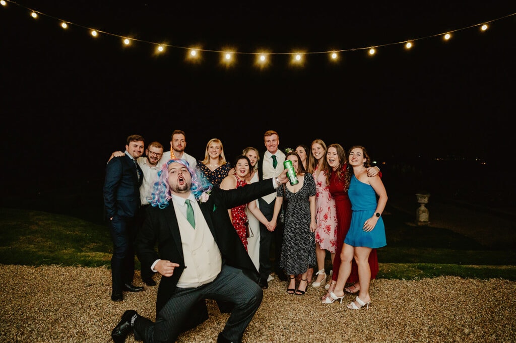 A group of people pose for a photo at night under string lights. One person in the foreground is making a playful gesture and wearing a colorful wig.