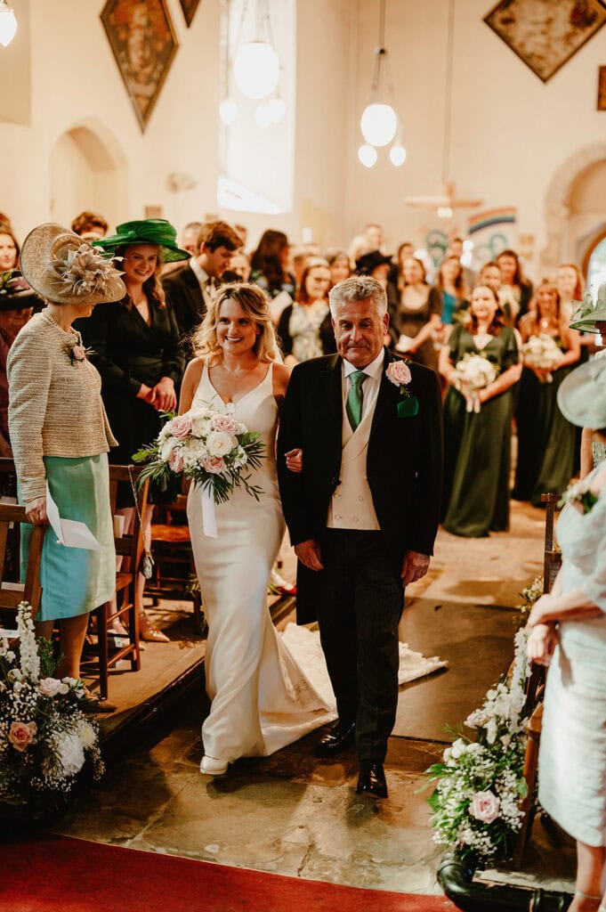 A bride in a white gown walks down the aisle with an older man in a suit, surrounded by guests dressed in formal attire inside a decorated venue.