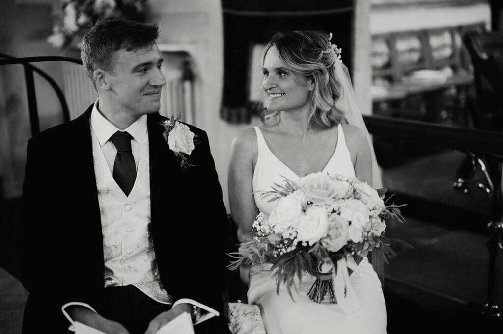 A bride and groom sit together, dressed in wedding attire. The bride holds a bouquet of flowers and the couple looks at each other, smiling. The background includes chairs and a draped cloth.