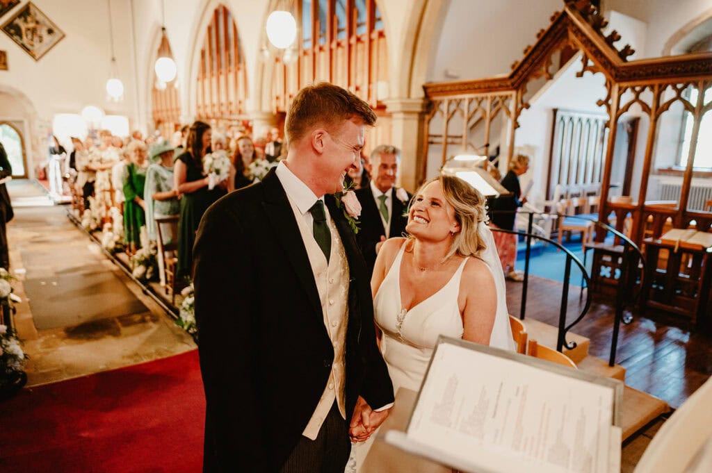 A couple stands at the altar, smiling at each other during their wedding ceremony in a church with guests seated and a lit aisle.