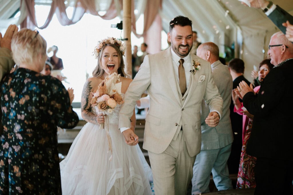 A bride and groom, dressed in wedding attire, walk down the aisle hand in hand, smiling and surrounded by applauding guests in a decorated venue.