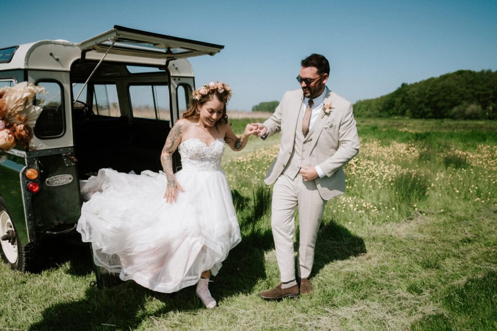 A bride and groom in wedding attire step out of a vehicle into a grassy field on a sunny day. The bride holds her dress, and the groom assists her, both smiling.