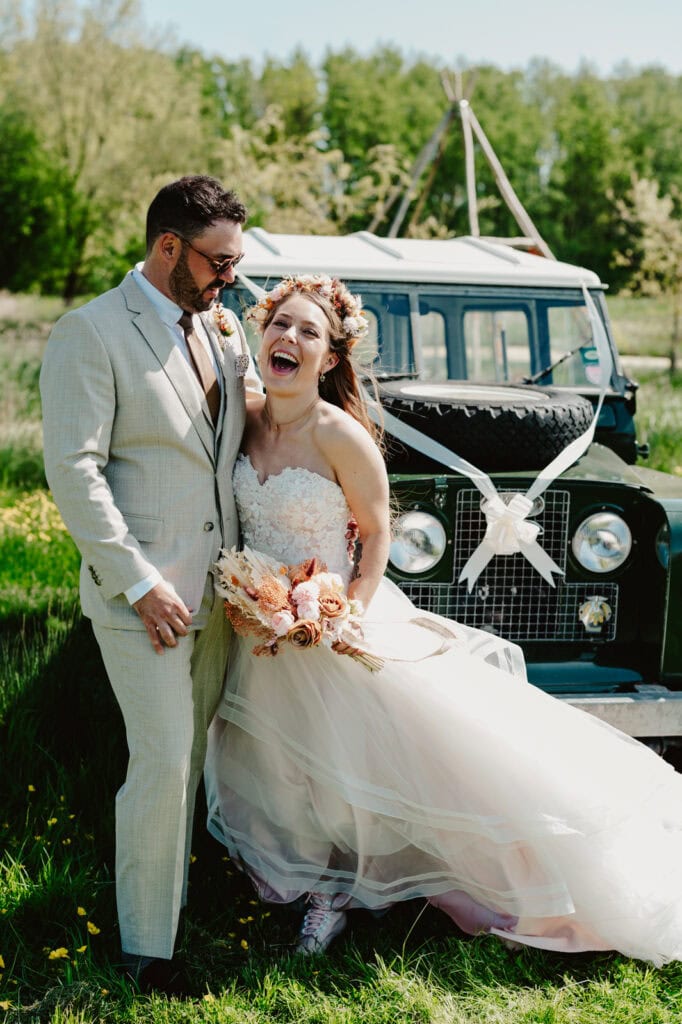 A bride and groom stand smiling and laughing in front of a decorated vintage off-road vehicle, with greenery and trees in the background. The bride holds a bouquet and wears a floral headpiece.