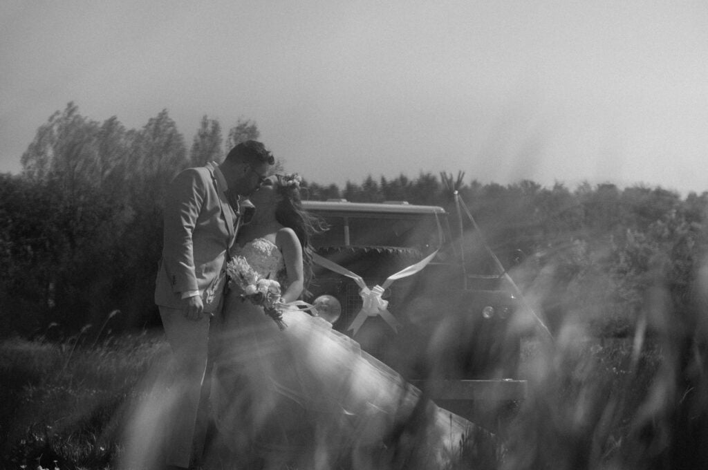 A bride and groom share a kiss outdoors next to a decorated vehicle, surrounded by tall grass and trees, in black and white.