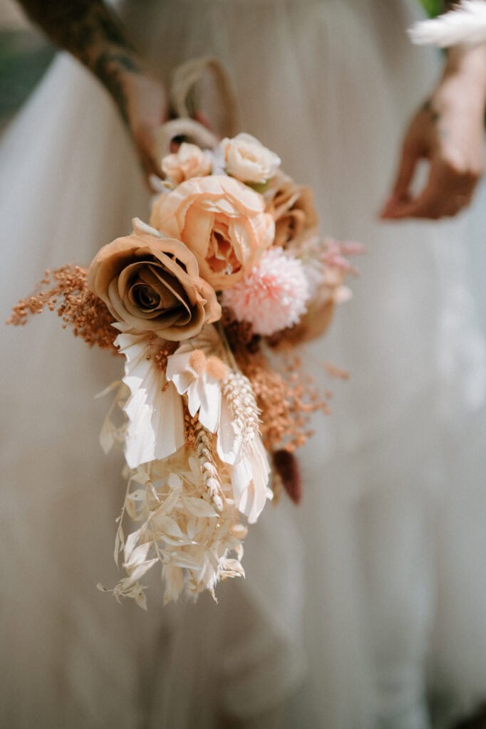 Close-up of a person holding a wedding bouquet with light brown roses, peach-colored flowers, and dried foliage. The person is wearing a white dress.