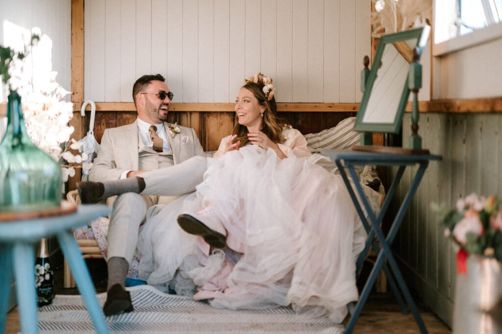 A bride in a flowing white dress sits relaxed next to a groom in a light gray suit and sunglasses. They're smiling and seated in a cozy, rustic setting at Wilderness Weddings Kent, with soft lighting, surrounded by wooden decor and flowers—capturing the essence of wedding photography perfection. Image by Pearce Wedding Photography.