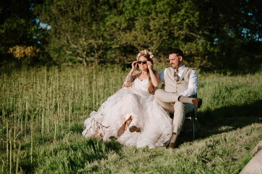 A bride and groom sit on a chair in a grassy field. The bride wears sunglasses and a flower crown, while the groom is in a beige suit. Trees are visible in the background.