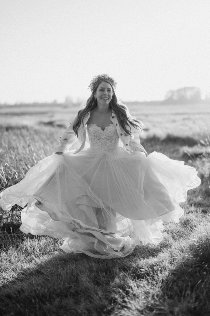 A bride in a field wears a flowing wedding gown with a lace bodice and a floral headpiece, holding her dress as she smiles and walks.