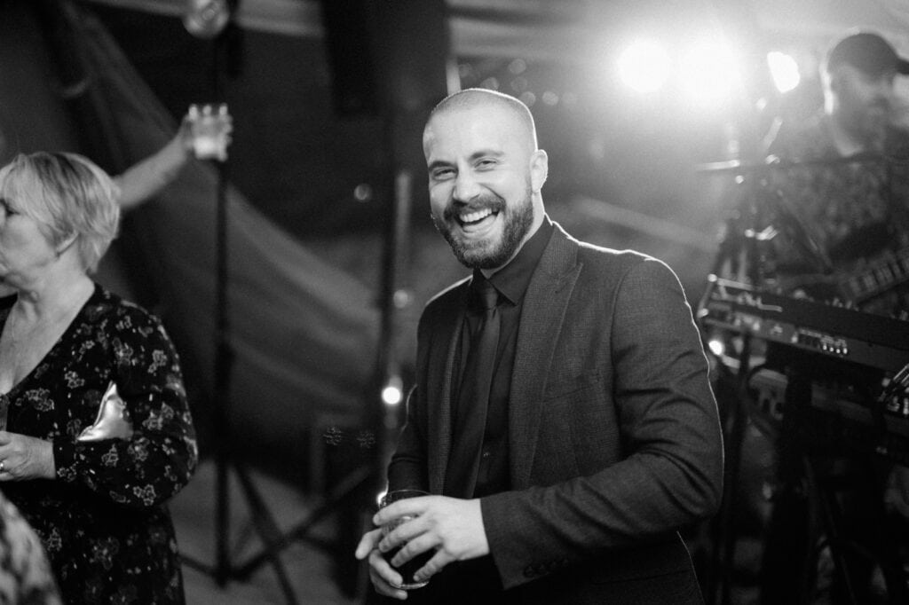 A man in a suit and tie smiles while holding a drink at an event. Another person is partially visible nearby and a band performs in the background under bright lights.