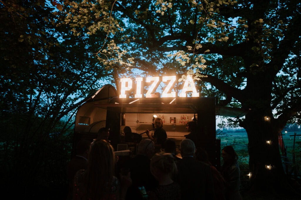 A lit-up "PIZZA" sign atop a food truck at night, surrounded by people and illuminated trees.