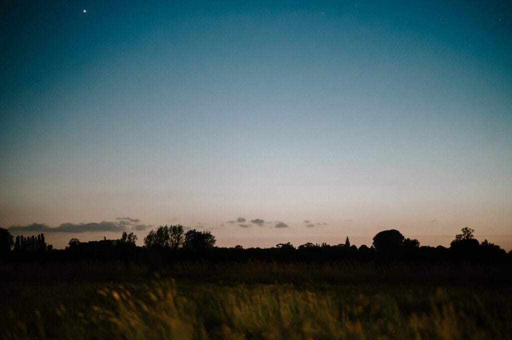 A dusk sky with a gradient from dark blue to pale orange is seen above a silhouetted landscape with trees and bushes.