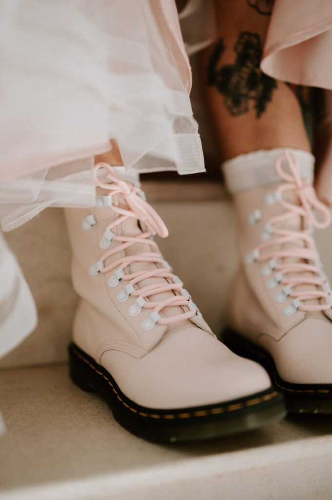Close-up of a person wearing light pink combat-style boots with white laces and a black sole. A tattoo is visible on their leg under the lightly ruffled hem of a garment.