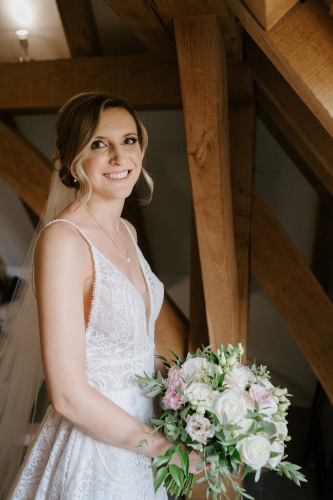 A bride in a white lace wedding gown stands indoors at The Old Kent Barn, smiling at the camera. She holds a bouquet of white and pink flowers with greenery. Her hair is styled in a loose updo, and a veil drapes over her shoulders. Wooden beams are visible in the background of this charming wedding scene. Image by Pearce Wedding Photography.