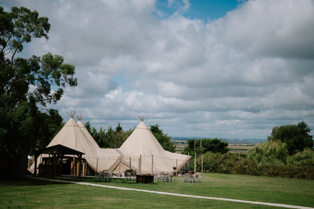 Outdoor wedding venue in Kent featuring large beige tents on a grassy field under a partly cloudy sky. Trees and open landscape add to the serene backdrop, perfect for your special day.