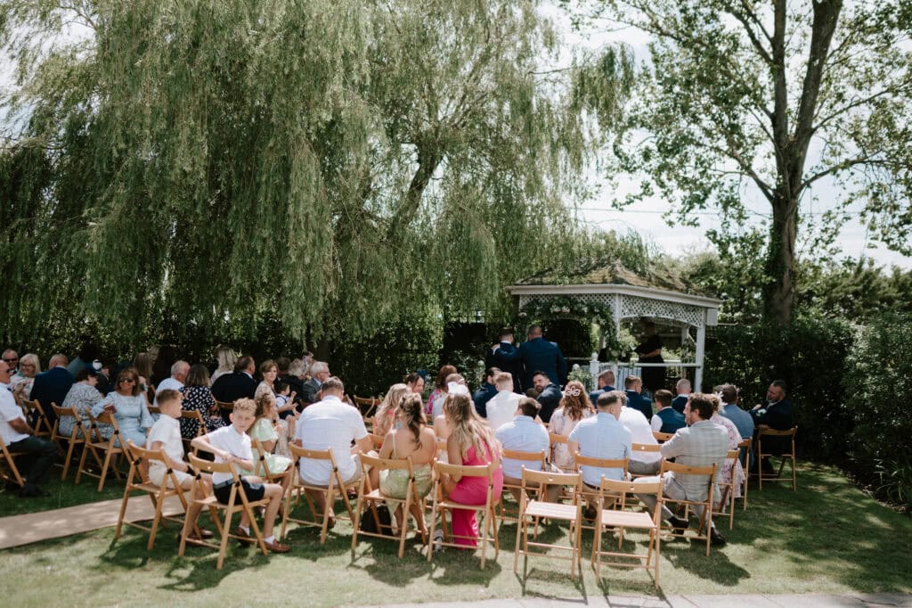 Outdoor wedding ceremony with guests seated on wooden chairs facing a beautifully decorated gazebo under the shade of trees. The sunny day in Kent highlights the enchanting setup, perfect for a romantic wedding.
