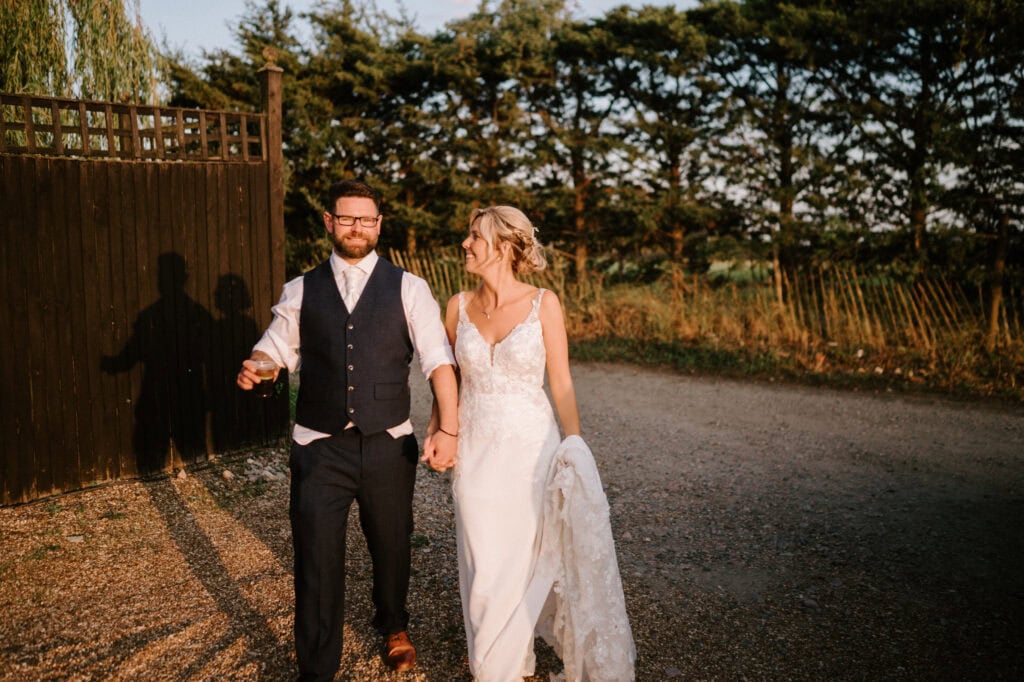 A couple, dressed in formal attire, walks hand in hand on a gravel path at a Kent wedding, with trees and a wooden fence in the background. The man holds a drink in his left hand.