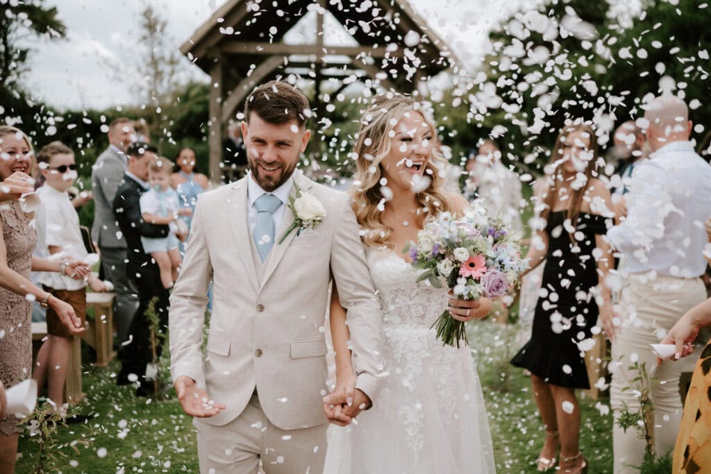 A bride and groom walk hand-in-hand down the outdoor aisle at Reach Court Farm during their charming Kent wedding, as guests throw white flower petals. The bride holds a bouquet of flowers, and both are smiling. A wooden structure is visible in the background.