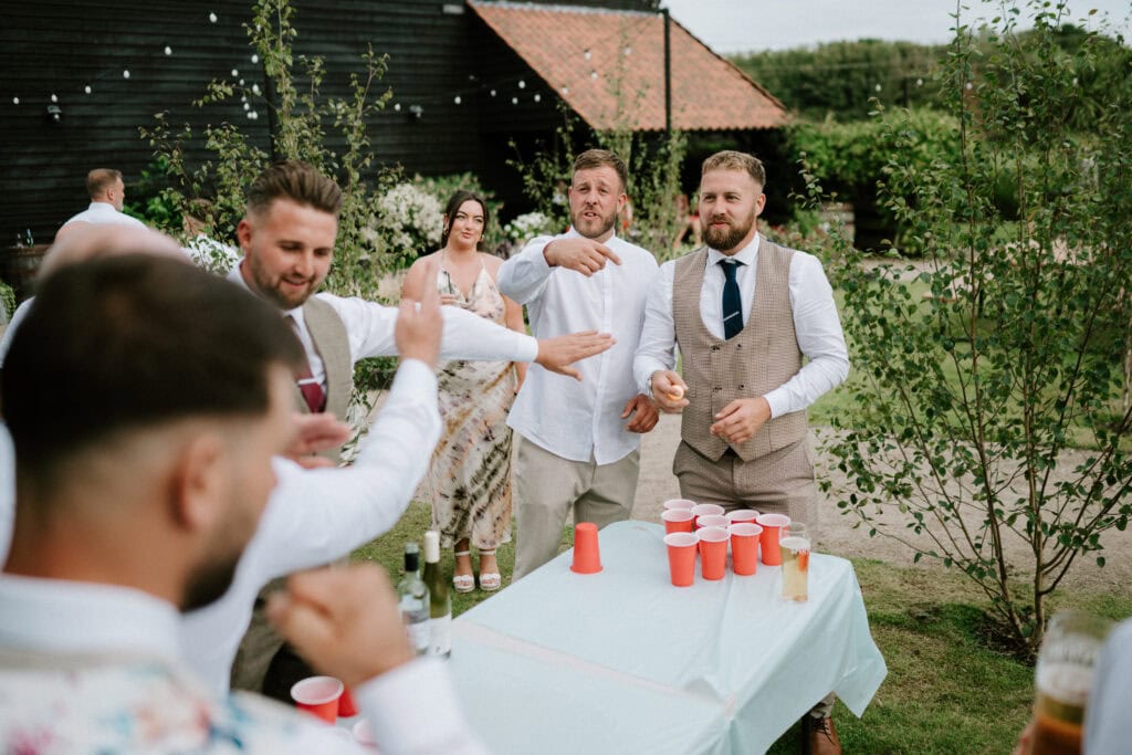 A group of people are playing a game of beer pong outdoors at Reach Court Farm. Some are in suits and one woman is in a floral dress. Red cups are on a table, and greenery surrounds the area, making it feel like the perfect setting for a Kent wedding celebration.