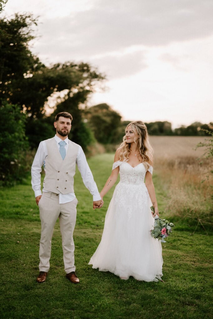 A bride and groom stand holding hands outdoors, dressed in formal wedding attire at the beautiful Reach Court Farm, Kent. The bride wears a white gown and holds a bouquet, while the groom sports a light vest and tie with a white shirt.