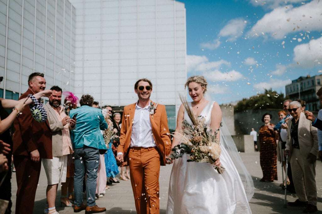 A bride and groom smile and walk down an outdoor aisle at their wedding in Margate, as guests throw confetti. The groom wears an orange suit, and the bride is in a white dress, holding a bouquet. The Turner Contemporary building serves as a striking backdrop.