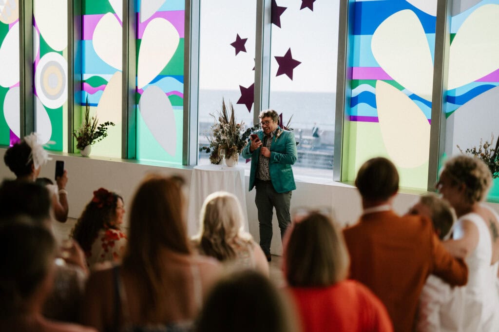 A person in a teal blazer speaks to a crowd at Turner Contemporary in Margate, standing before vibrant, colorful windows decorated with abstract shapes and star motifs, with the sea visible outside.