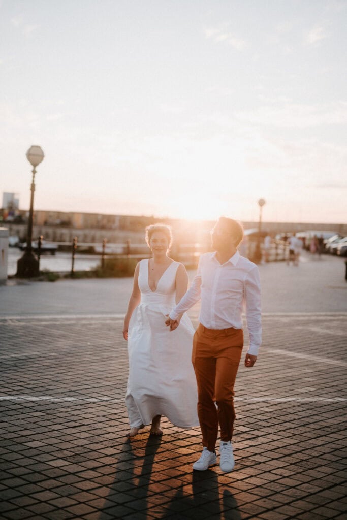 A couple walks hand in hand on a paved waterfront path at sunset, near Margate's Turner Contemporary. The person in front is wearing a white dress, and the other person is dressed in a white shirt and brown pants, reminiscent of a serene post-wedding stroll.