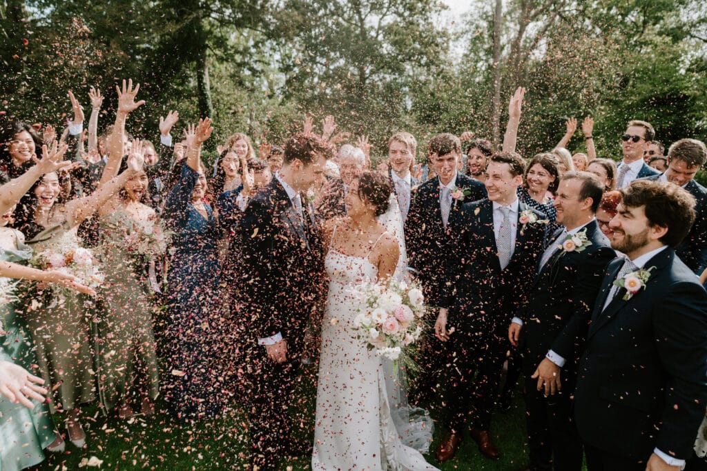 A newlywed couple stands in the center of a group of people who are showering them with confetti, while everyone celebrates outdoors in a garden setting. The joyous occasion is perfectly captured by the talented wedding photographer at this enchanting Port Lympne wedding.