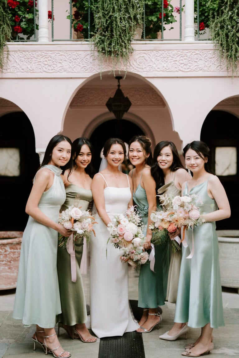 A bride in a white dress stands with five bridesmaids in green dresses, smiling and holding bouquets, at a Port Lympne wedding. The outdoor setting features pink arches and lush greenery in the background, captured beautifully by their photographer.