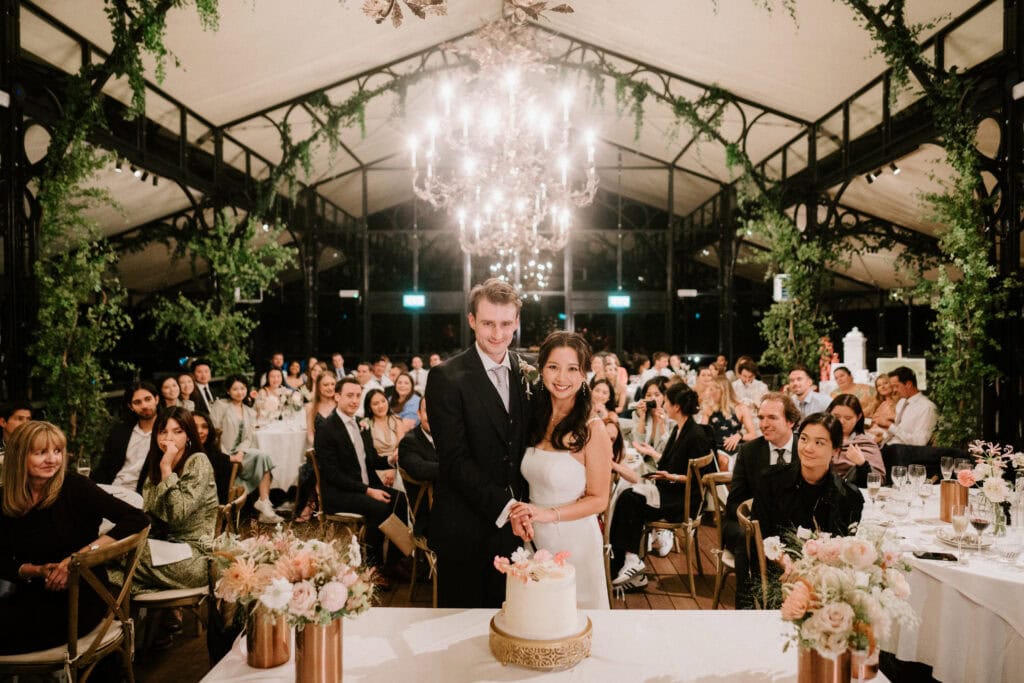 A bride and groom are cutting a wedding cake in a decorated reception hall, surrounded by guests seated at tables adorned with floral arrangements. A photographer captures the moment as a chandelier hangs above them, immortalizing their beautiful Port Lympne wedding day.