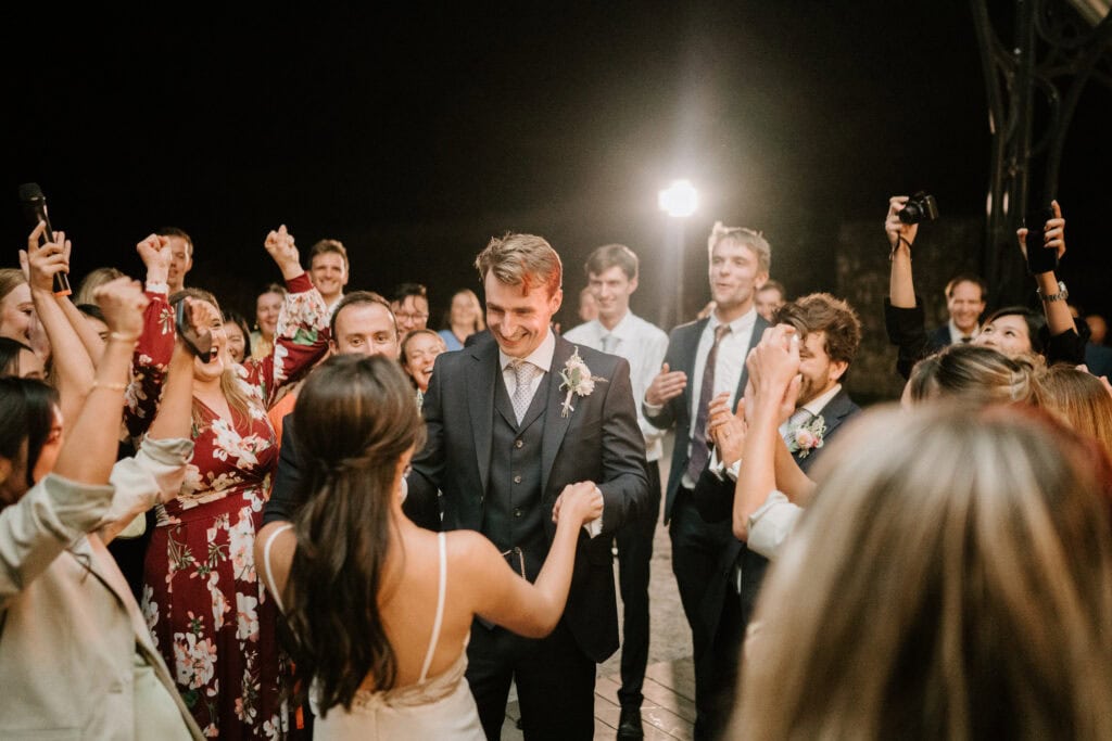 A couple in formal attire, surrounded by cheering guests at a picturesque Port Lympne wedding, appear to be celebrating at a nighttime event. The man is smiling while holding the woman's hand, perfectly captured by the photographer.