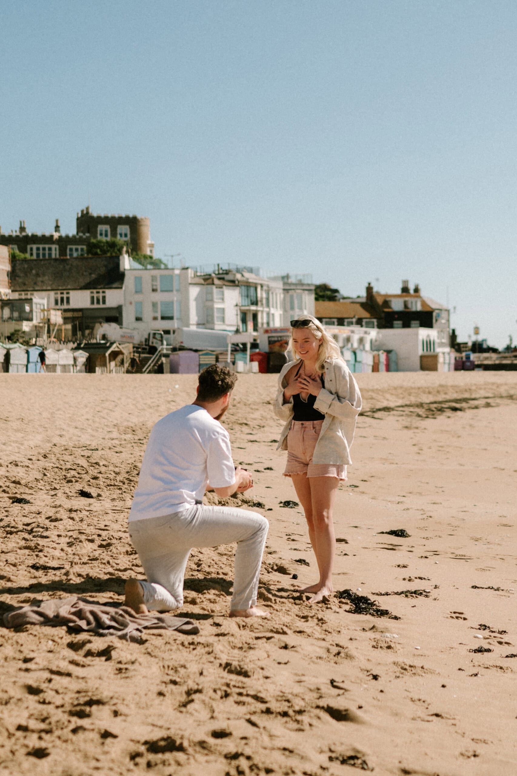 Beach proposal with surprised woman in front of seaside buildings.
