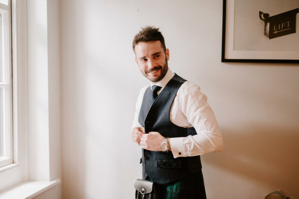 A man with short dark hair and a beard stands in a well-lit room near a window, smiling and buttoning his dark vest over a white shirt. A kilt is partially visible. A framed picture hangs on the wall behind him, capturing this moment perfectly—as if seen through the eyes of a Bethnal Green wedding photographer. Image by Pearce Wedding Photography.