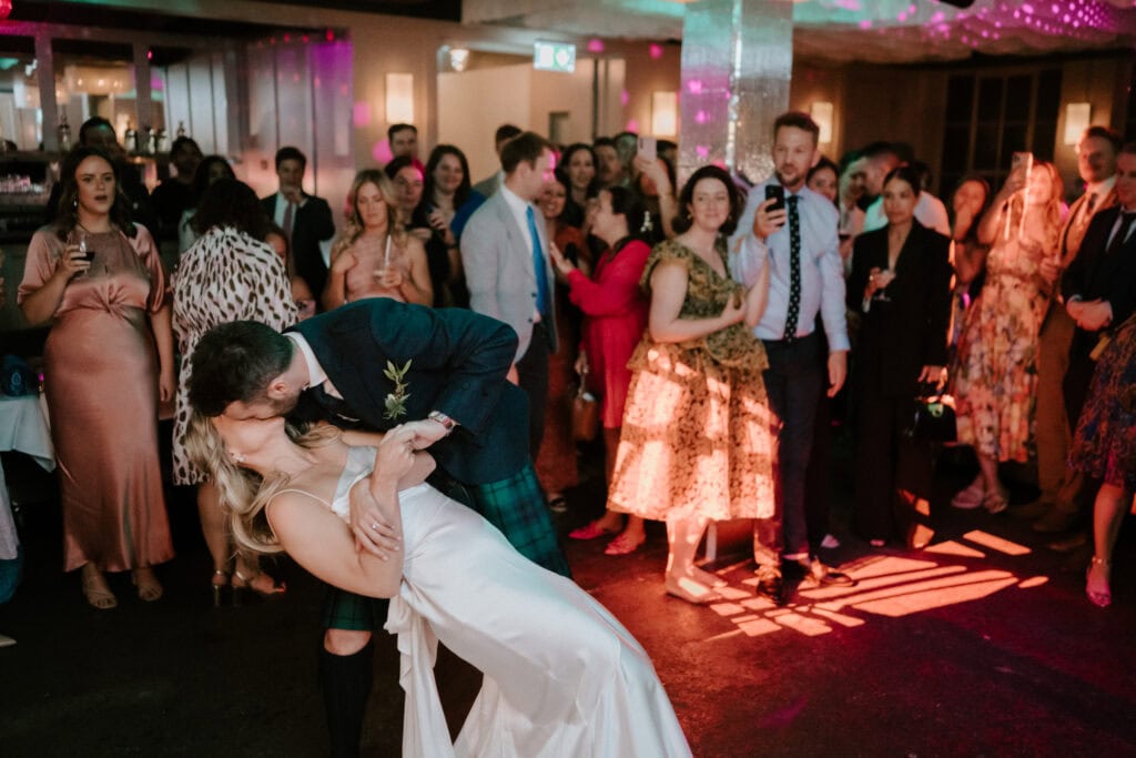 A couple shares a kiss while dancing at a lively wedding reception. The groom dips the bride as they kiss, surrounded by elegantly dressed guests who watch with smiles, some capturing the moment on their phones. The room is decorated with warm, colorful lighting—a scene beautifully immortalized by a Bethnal Green wedding photographer. Image by Pearce Wedding Photography.