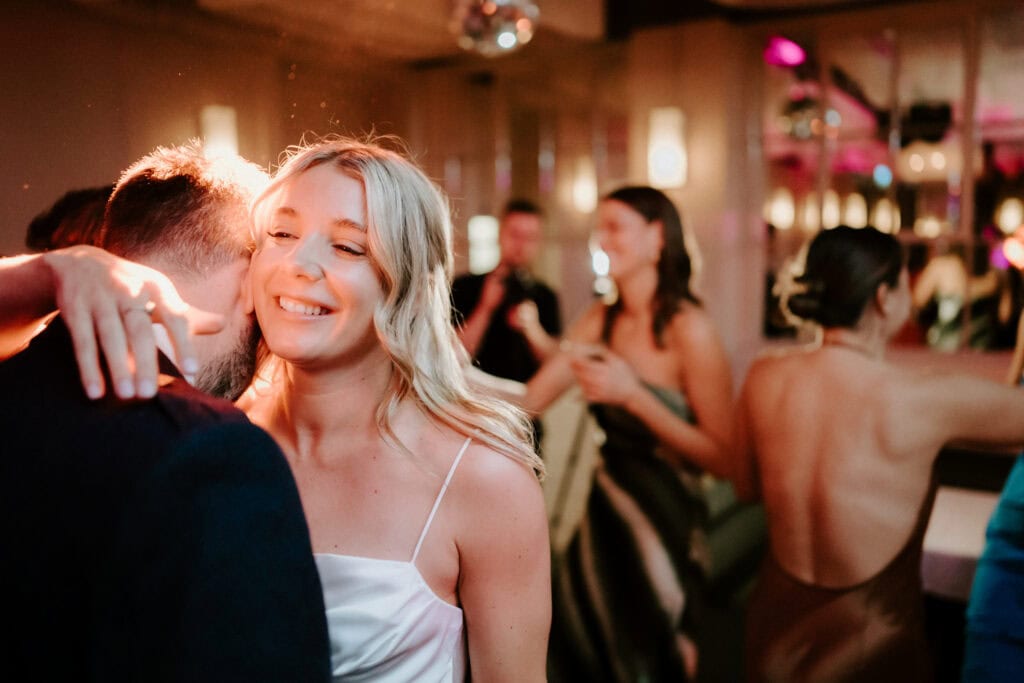 A woman with blonde hair smiles and hugs a man with a beard in a warmly lit room. Other people are in the background, some dancing and others conversing. The atmosphere suggests a festive or celebratory event, beautifully captured by the Bethnal Green wedding photographer. Image by Pearce Wedding Photography.