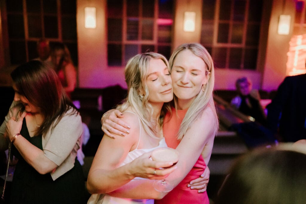Two women are embracing warmly while one holds a drink. Both have light-colored hair and are smiling with their eyes closed. The setting appears to be indoors with warm lighting, and other people can be seen in the background—captured perfectly by a Bethnal Green wedding photographer. Image by Pearce Wedding Photography.
