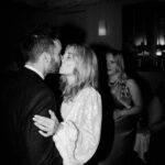 A couple is captured in a black-and-white photo, sharing a kiss. The woman is wearing a shimmering dress, while the man is dressed in a suit. They are surrounded by other people in what appears to be a warmly lit indoor setting with soft decor, expertly framed by their Bethnal Green wedding photographer. Image by Pearce Wedding Photography.