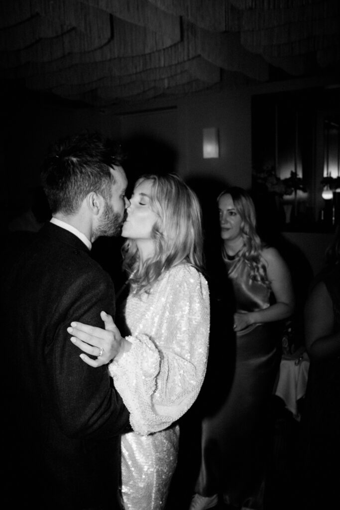 A couple is captured in a black-and-white photo, sharing a kiss. The woman is wearing a shimmering dress, while the man is dressed in a suit. They are surrounded by other people in what appears to be a warmly lit indoor setting with soft decor, expertly framed by their Bethnal Green wedding photographer. Image by Pearce Wedding Photography.
