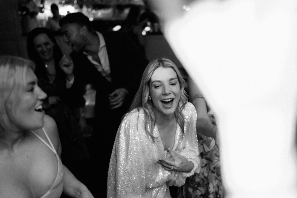 A black and white photo of several people dancing at a party or wedding reception. A woman in the center is laughing and has her hand on her chest, captured perfectly by a Bethnal Green wedding photographer. Other people around her are also enjoying and dancing. The atmosphere is lively and cheerful. Image by Pearce Wedding Photography.