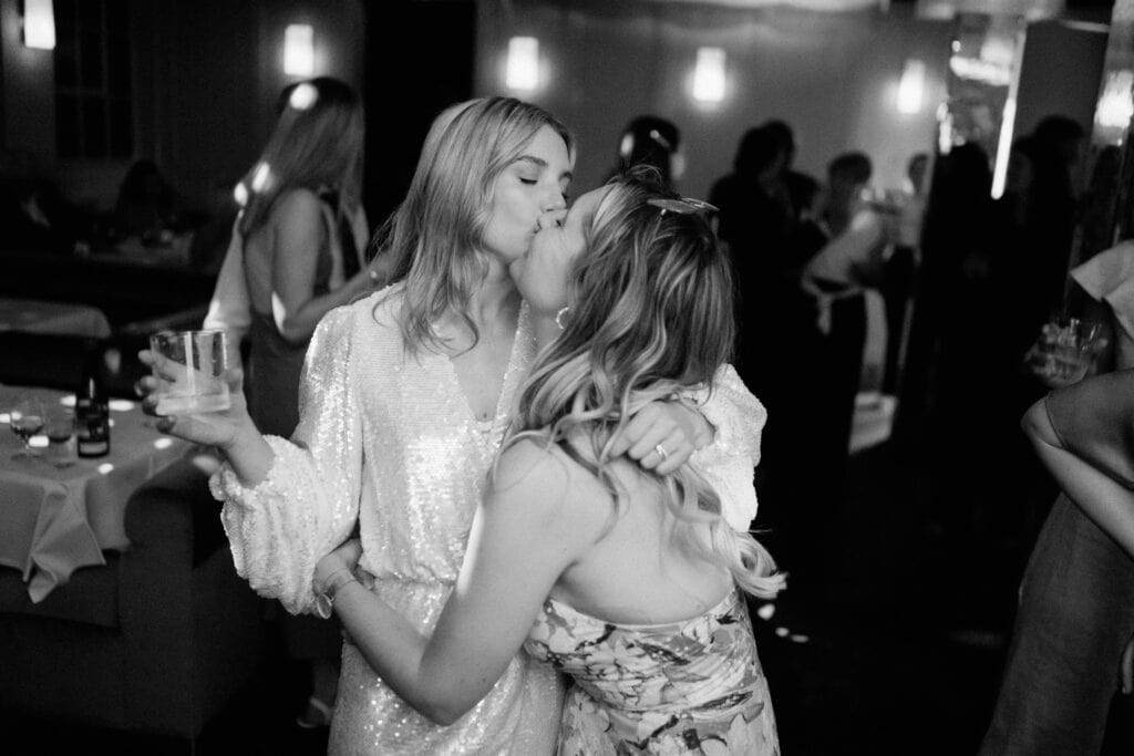 Black and white image of two women kissing at an indoor event or party. One woman is wearing a sparkling dress and holding a drink, while the other has her arm around her and is also holding a drink. People are visible in the blurred background, captured beautifully by a Bethnal Green wedding photographer. Image by Pearce Wedding Photography.
