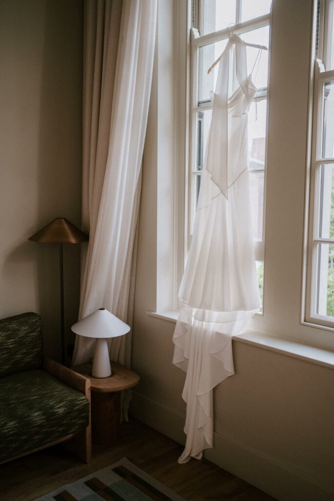 A long, elegant white dress hangs from a window, draping gracefully toward the floor. The setting includes a green upholstered chair, a wooden side table, a modern white lamp, and a floor lamp, all illuminated by natural light streaming through the window—perfect for any Bethnal Green wedding photographer's shot. Image by Pearce Wedding Photography.