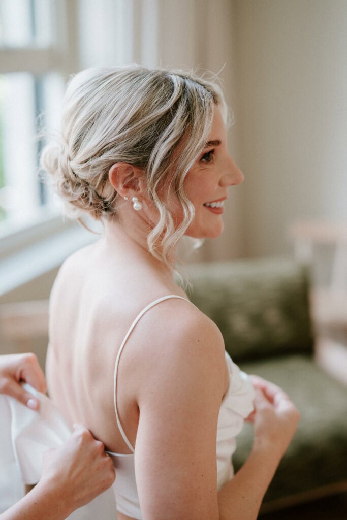 A woman with light blonde hair styled in an updo smiles while looking to her right. She is wearing a white sleeveless dress, with someone adjusting it from behind. Captured by a Bethnal Green wedding photographer, the blurred background shows a window and green cushioned furniture. Image by Pearce Wedding Photography.
