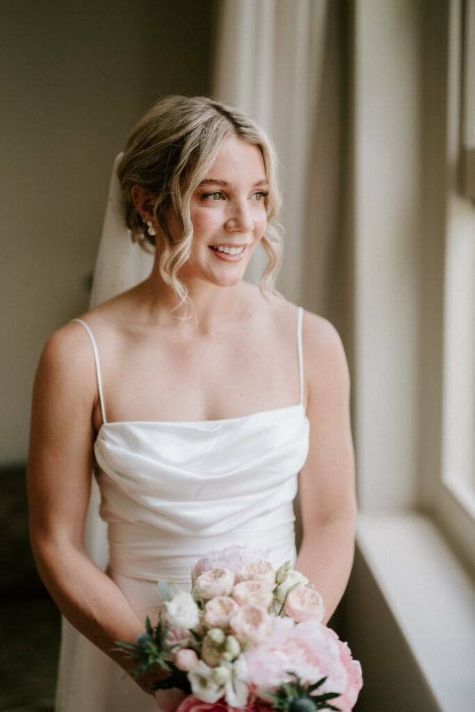 A bride with wavy blonde hair and a veil smiles while standing near a window. She wears a sleeveless white wedding dress and holds a bouquet of pale pink and white flowers. Captured by the talented Bethnal Green wedding photographer, the soft light from the window illuminates her face and dress. Image by Pearce Wedding Photography.