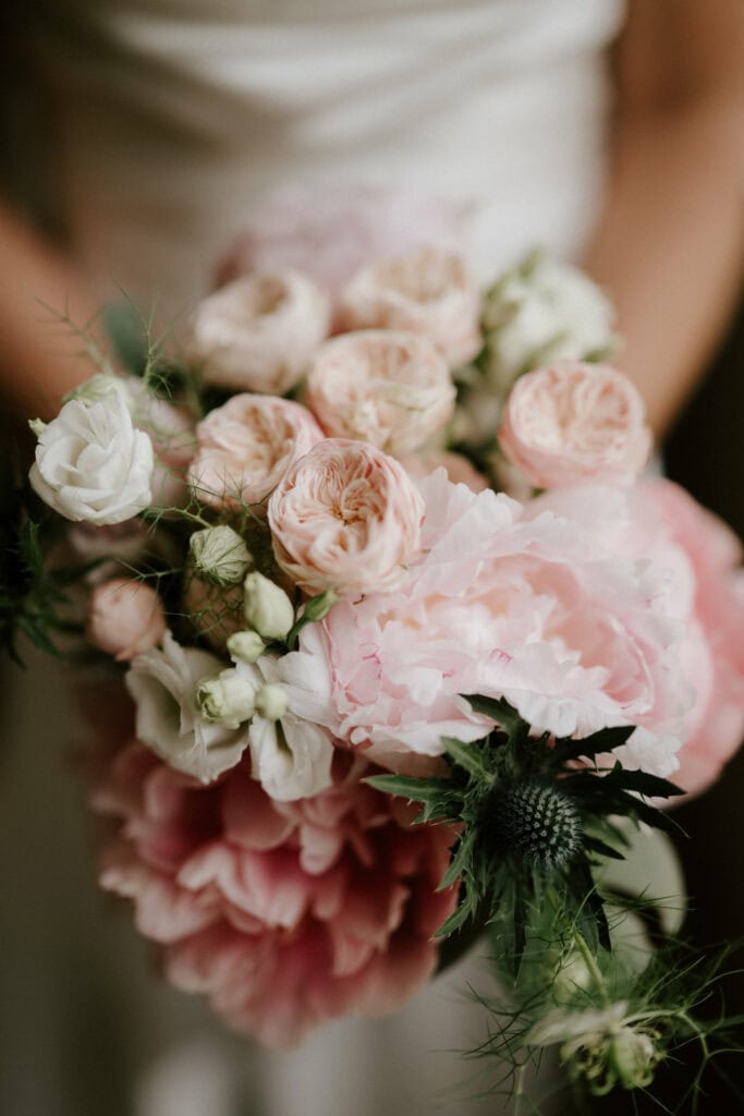 A close-up of a person holding a bouquet of flowers. The arrangement features a mix of light pink roses, white flowers, and greenery, with delicate textures and soft shades, creating an elegant and romantic appearance—perfect for capturing by a Bethnal Green wedding photographer. Image by Pearce Wedding Photography.