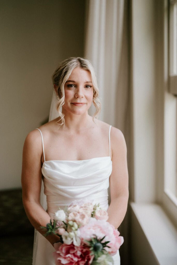 A bride in a white dress stands near a window holding a bouquet of pink and white flowers. She has blonde hair styled in loose curls and a gentle expression, perfectly captured by her Bethnal Green wedding photographer. The room appears soft-lit with neutral tones. Image by Pearce Wedding Photography.